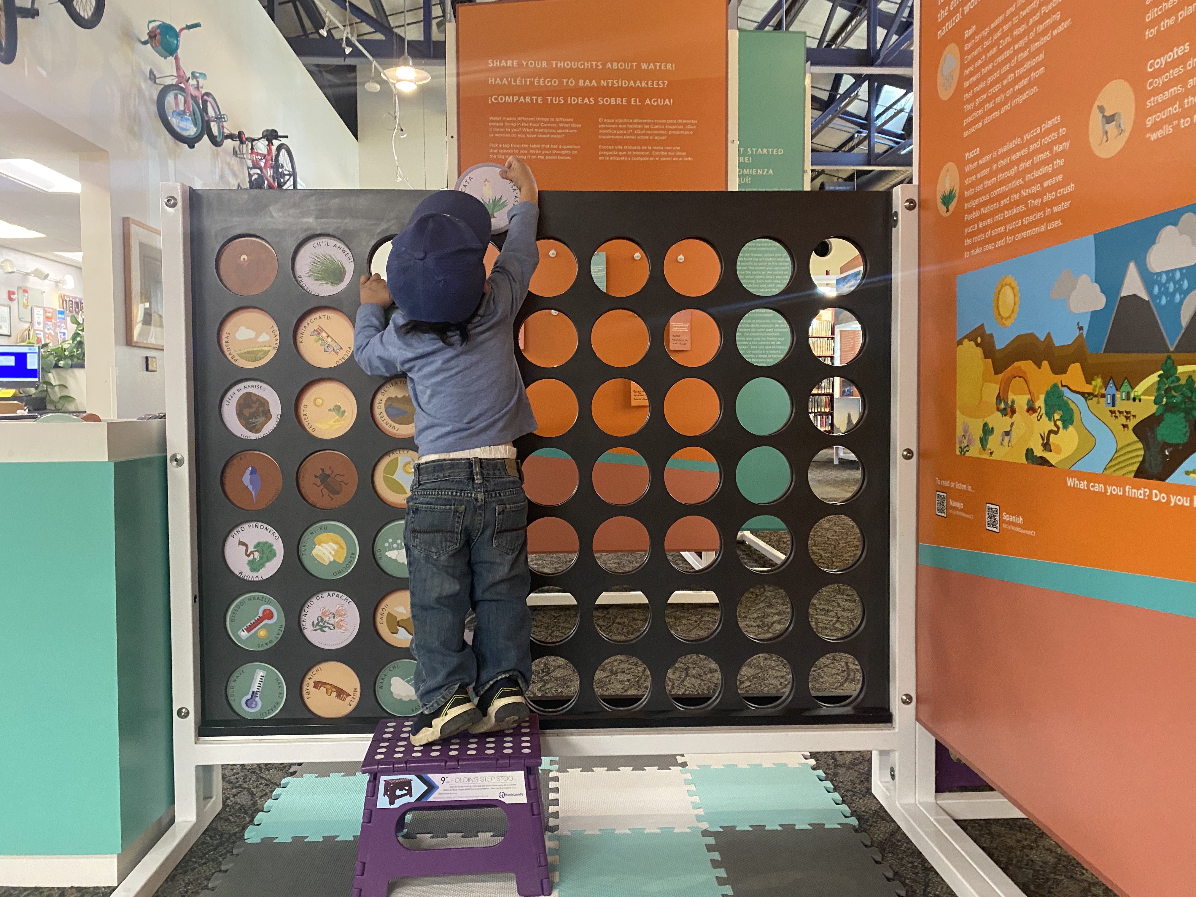 A young child stands on a step stool and reaches above the Connect 4 board to insert a game token.